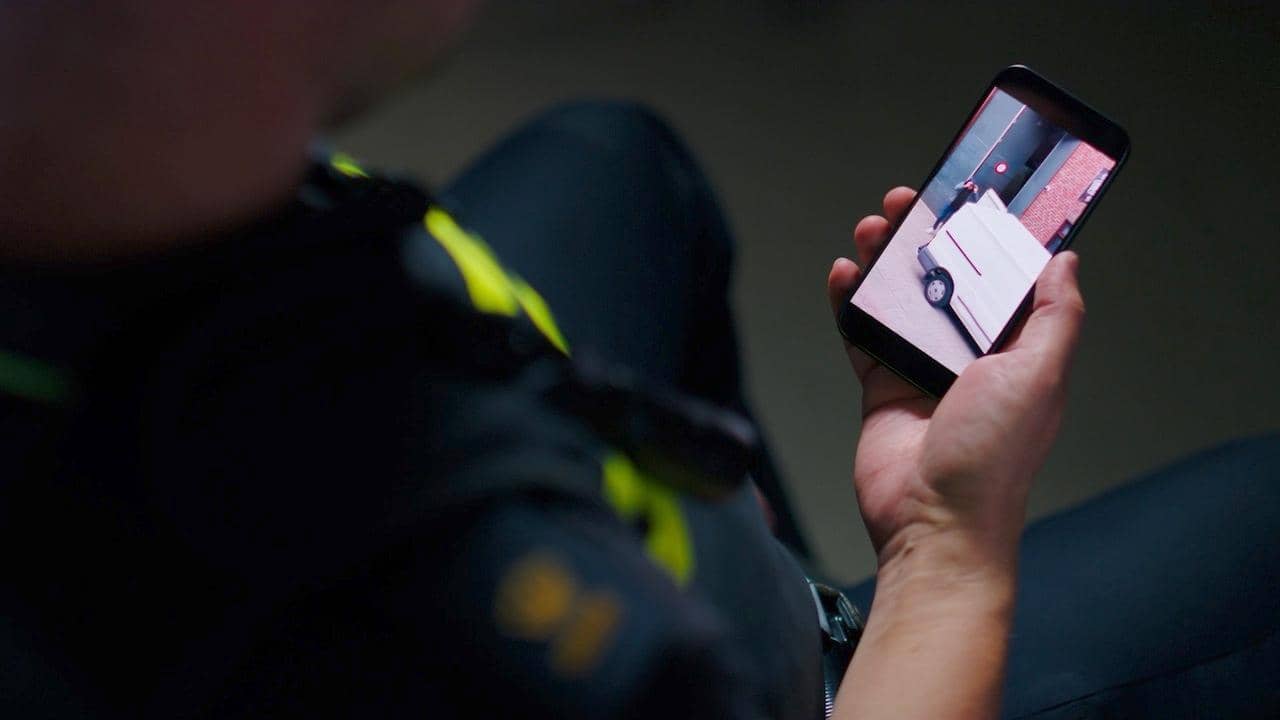Politieagent houdt een mobiel scherm vast waarop te zien is hoe een wit busje wordt uitegeladen.