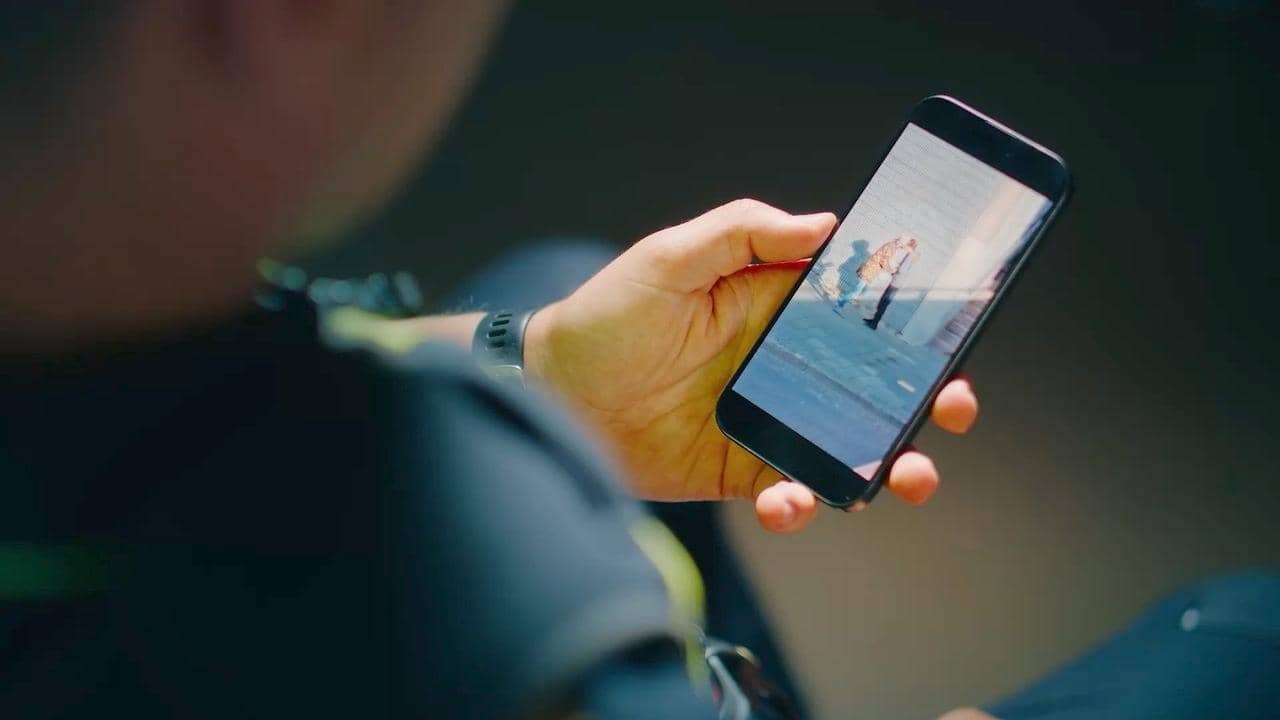 Een agent houdt een telefoonscherm vast met daarop een beeld van twee mensen die elkaar iets overhandigen.